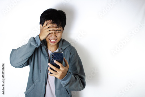 Asian teenager in white t-shirt and blue hoodie, posing angrily and covering his face while looking at his cellphone