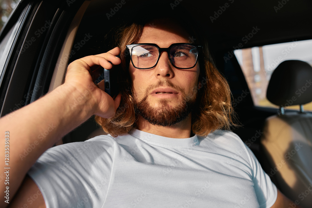 Young man with glasses making a phone call in a car, looking thoughtful and engaged Young man with glasses making a phone call in a car, looking thoughtful and engaged