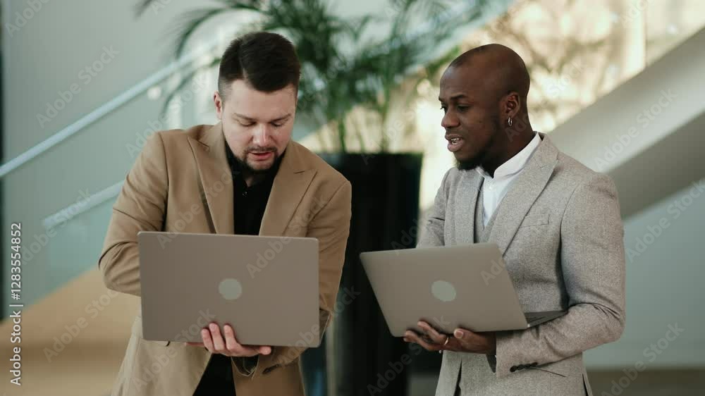 Two business men, an African American and a European, discuss a project before a conference in the hallway of a modern office building. Concept professional atmosphere, collaboration, and technology. Two business men, an African American and a European, discuss a project before a conference in the hallway of a modern office building. Concept professional atmosphere, collaboration, and technology.
