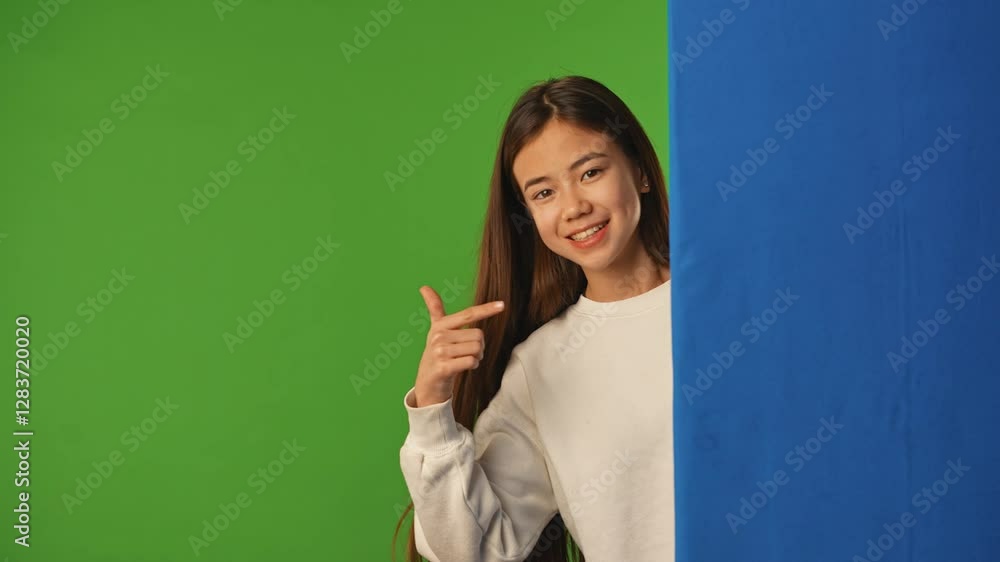 Asian girl child peeks out and waves hand, pointing at blue space, positive expression, isolated on chroma key green screen background mockup. Asian girl child peeks out and waves hand, pointing at blue space, positive expression, isolated on chroma key green screen background mockup.