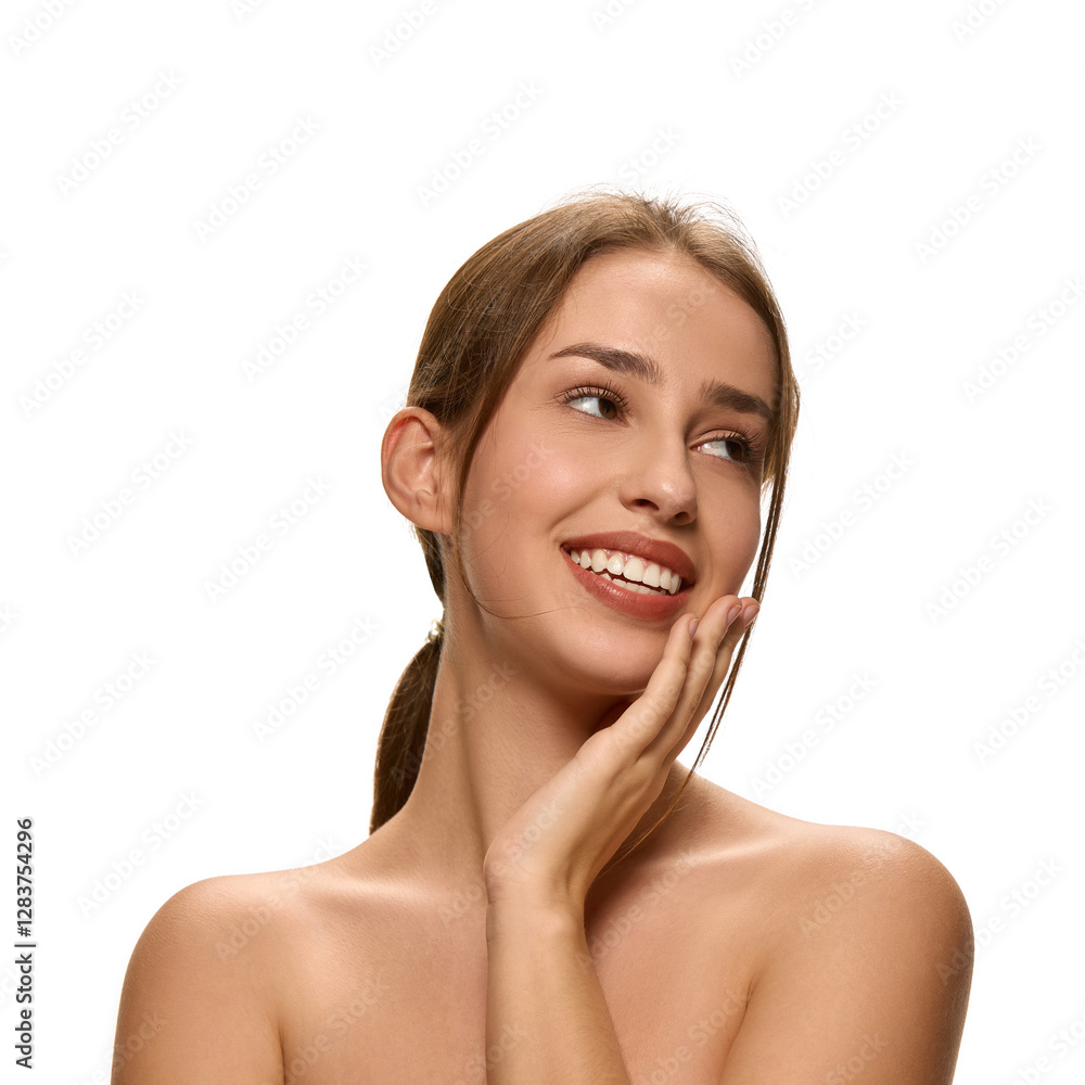 Joyful young woman smiling and touching cheek while looking sideways against white studio background. Concept of beauty, self-care treatment, inner glow, confidence, cosmetic products. Joyful young woman smiling and touching cheek while looking sideways against white studio background. Concept of beauty, self-care treatment, inner glow, confidence, cosmetic products.