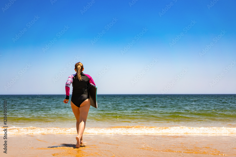 Fototapeta premium Water sports. Mature woman holding bodyboard running out of water on sunny beach. Back view