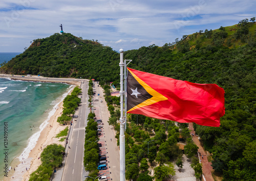 The national flag of Timor Leste flutters in the air with the Cristo Rei Beach and Statue in the background in Dili City, Timor Leste.
