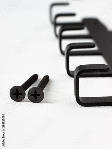 A close-up of two black cross-head screws lying on a white surface next to a metal wall hanger for hooks.
