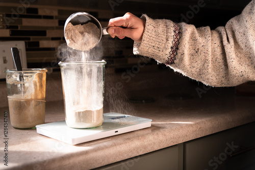 Person feeding sourdough starter and pouring flour into glass canister on a scale
