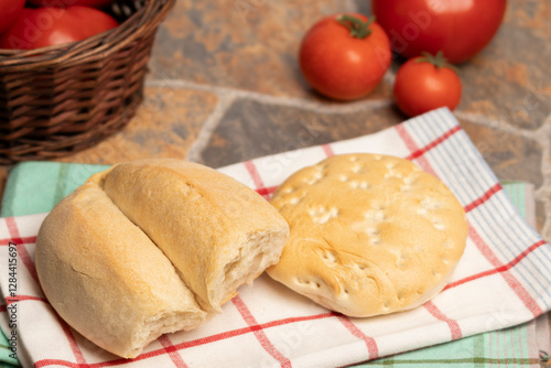 Un par de panes de trigo, una marraqueta y una hallula sobre un paño de cocina, al fondo un canasto con tomates y tomates rojos y maduros