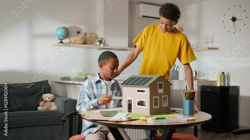 Younger brother is actively working on details of solar-powered model house as older brother joins, bringing his energy and ideas to project. Their seamless interaction reflects shared responsibility