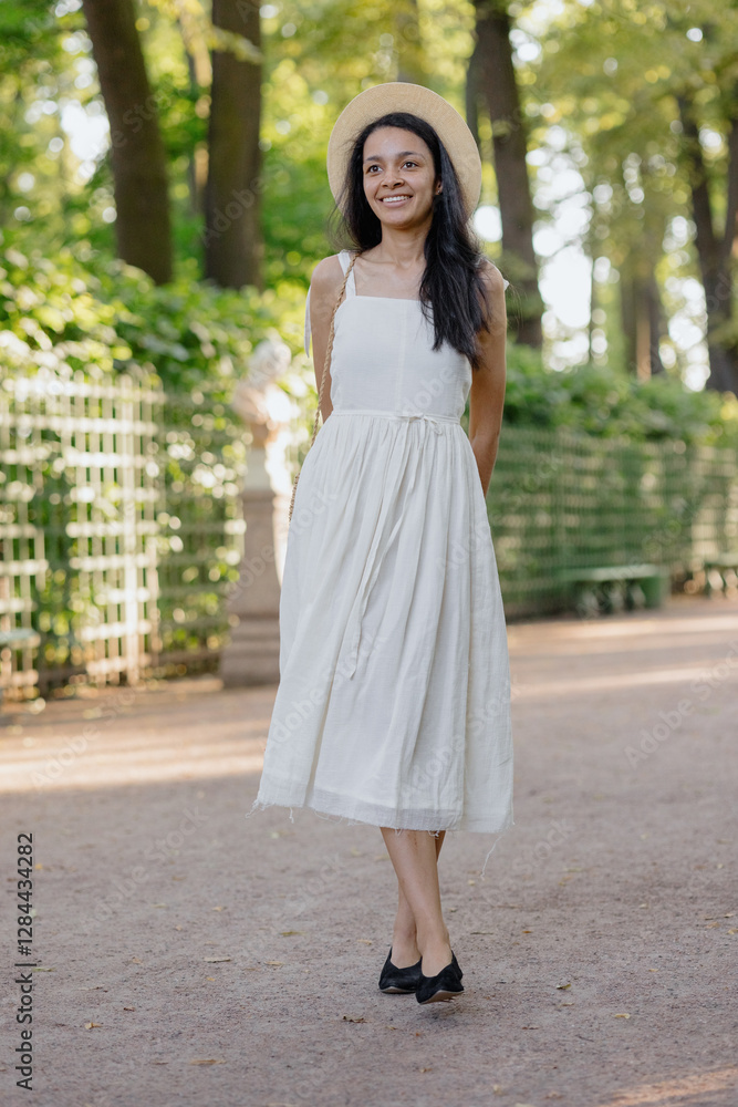 carefree African American woman in white dress and straw hat walks in park and poses against background of green hedge. daytime for relaxation. beautiful female fashion model, sunlight summer time carefree African American woman in white dress and straw hat walks in park and poses against background of green hedge. daytime for relaxation. beautiful female fashion model, sunlight summer time