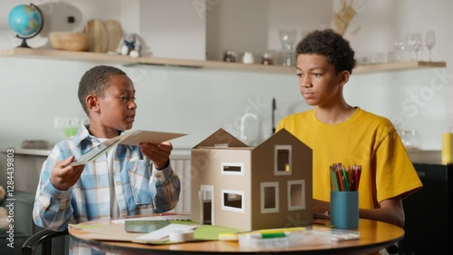 Both brothers work together to attach roof with solar panels to model house. This moment captures their cooperative effort, show problem-solving and teamwork in key parts of school science project