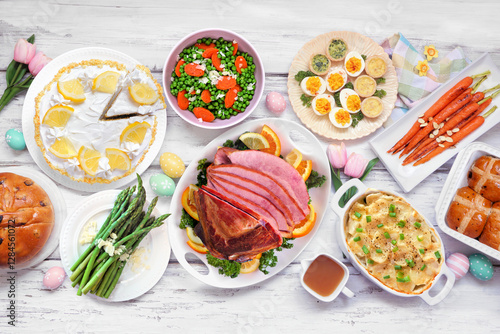 Classic Easter ham dinner. Overhead view table scene on a white wood background. Ham, scalloped potatoes, vegetable side dishes, eggs, breads and lemon cake dessert. photo