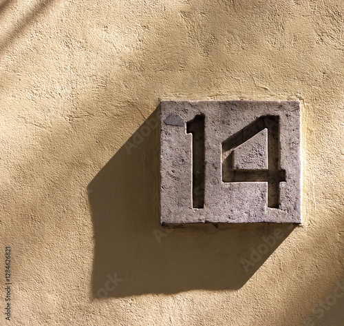 Street address number, number 14, inset into concret mounted on a yellow stucco wall.