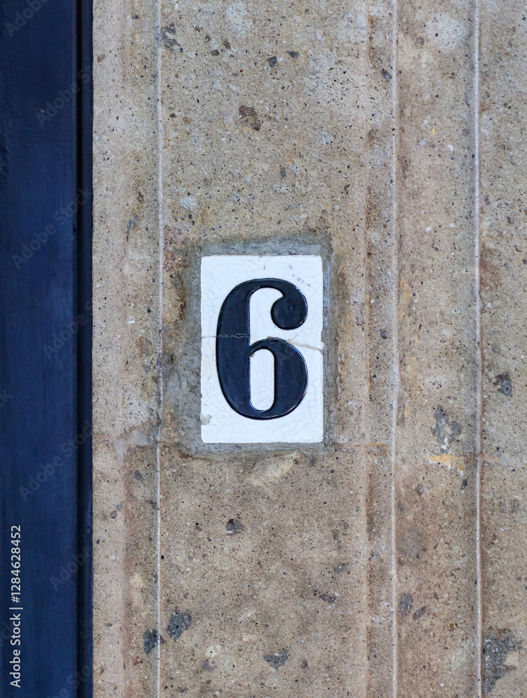 Street address number, number six (6). Cracked white tile with a dark blue number inset into a brown carved stone wall. Street address number, number six (6). Cracked white tile with a dark blue number inset into a brown carved stone wall.