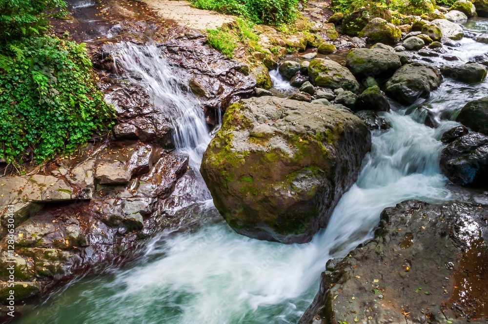 A small waterfall rushes over moss-covered boulders in a tranquil forest stream, creating a serene scene. A small waterfall rushes over moss-covered boulders in a tranquil forest stream, creating a serene scene.
