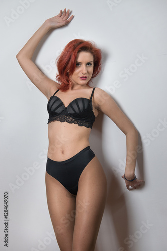 Obraz na płótnie Attractive red hair model with black lingerie standing against gray wall. Portrait of sensual long legs woman, indoor shot. Sensual redhead with perfect body posing provocatively with black bra w salonie