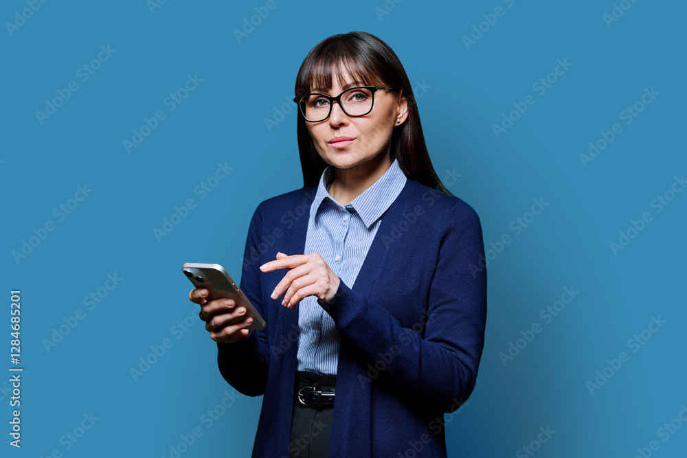 Middle aged businesswoman holding smartphone looking at camera on blue background Middle aged businesswoman holding smartphone looking at camera on blue background
