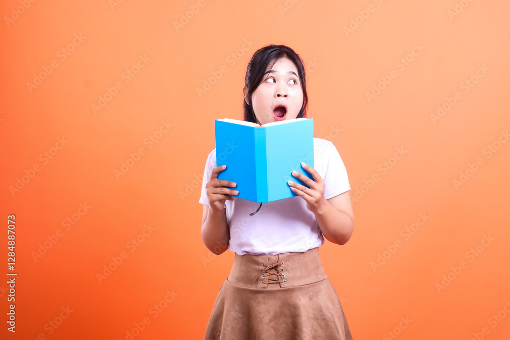 A person is holding a book with a surprised expression on her face against a solid orange background A person is holding a book with a surprised expression on her face against a solid orange background