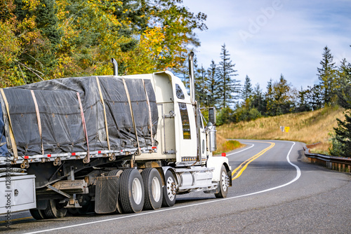 Industrial standard big rig white semi truck tractor with extended cab transporting covered and fastened cargo on flat bed semi trailer running on the winding mountain road with autumn trees