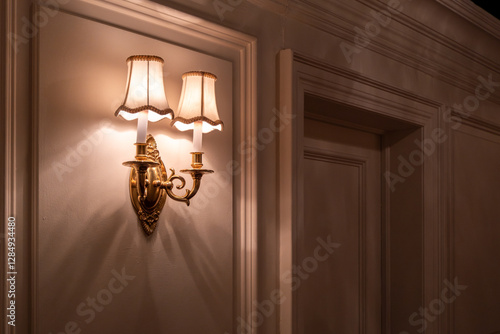 Elegant vintage wall sconce with dual fabric lampshades casts a warm, atmospheric glow on a classic paneled wall. Traditional brass light fixture creates dramatic shadows and ambiance in the hallway.