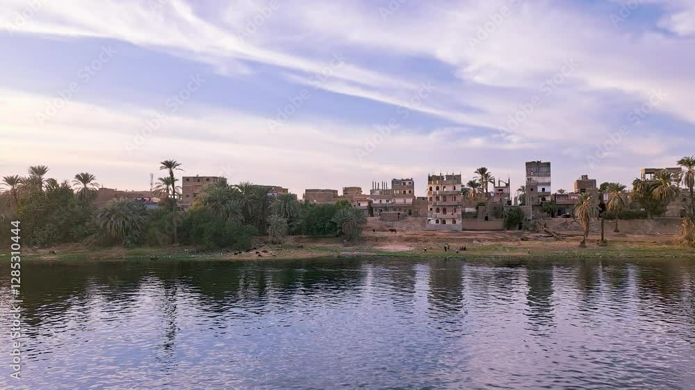 The riverbank is lined with lush green palm trees and colorful buildings. The calm blue water reflects the sky. The historical significance of the area for tourism, Luxor, Egypt. The riverbank is lined with lush green palm trees and colorful buildings. The calm blue water reflects the sky. The historical significance of the area for tourism, Luxor, Egypt.