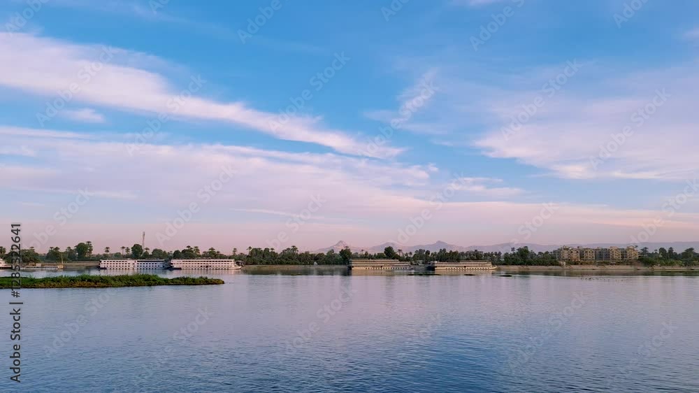 The riverbank is lush with green trees and palm trees. A small boat floats on the calm water. The traditional houses and serene atmosphere, Luxor, Egypt. The riverbank is lush with green trees and palm trees. A small boat floats on the calm water. The traditional houses and serene atmosphere, Luxor, Egypt.
