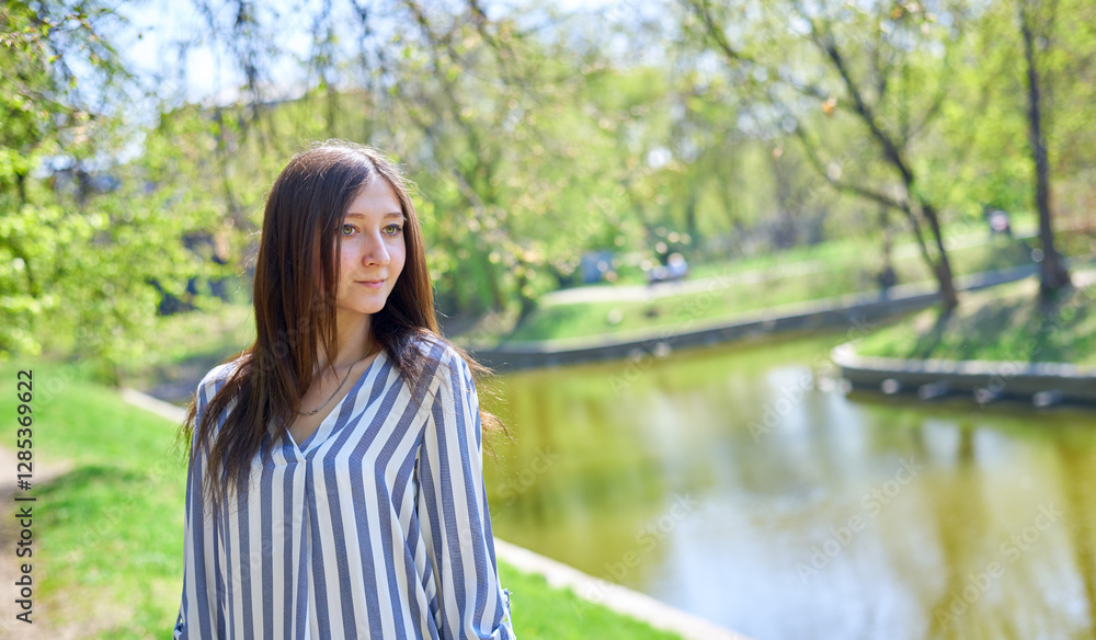 A woman stands in a park with trees in the background. She is wearing a striped dress and has her hand on her hip. A woman stands in a park with trees in the background. She is wearing a striped dress and has her hand on her hip.