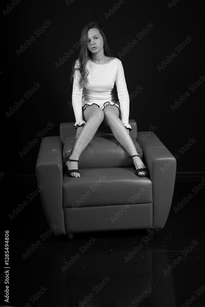 Fototapeta premium Long legged young woman in white dress sitting on a chair black and white low key portrait