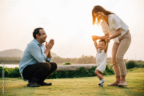 A smiling mother and father watching with joy as their baby takes his first steps in the park surrounded by nature at sunset time.
