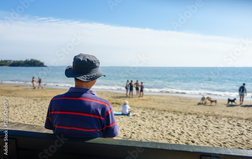 Obraz na płótnie Man sitting on the bench in summer. Unrecognizable people and dogs walking on Milford Beach. Auckland. w salonie