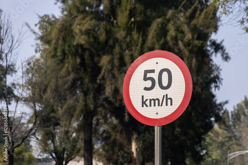 Fotografie 50 km per hour speed limit sign with a red border stands on a roadside, surrounded by trees and natural scenery in daylight
