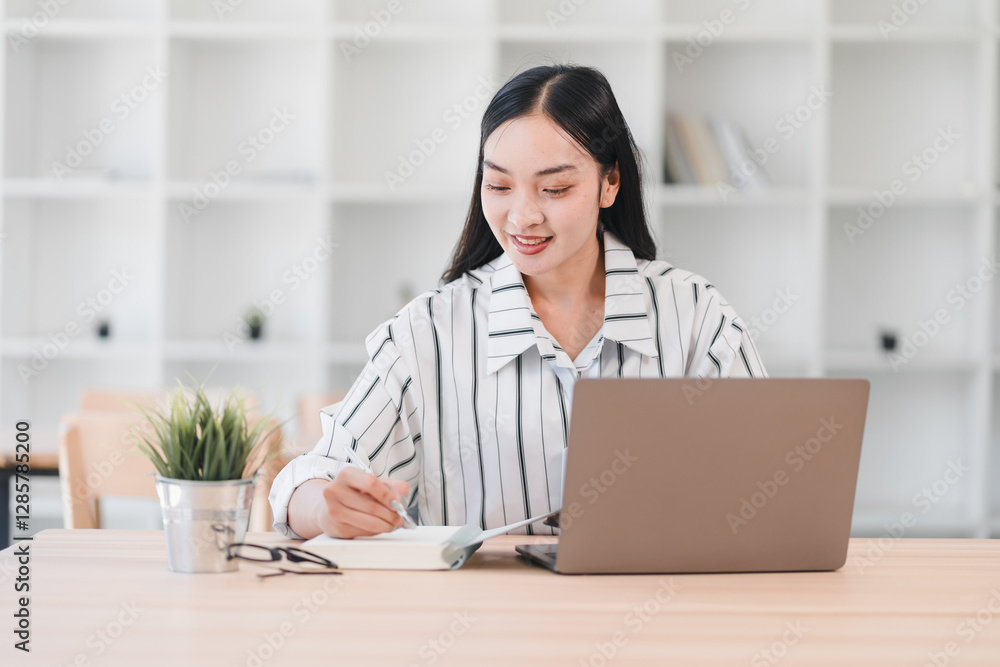 Woman working on laptop while taking notes in notebook, sitting at desk in bright office space. Woman working on laptop while taking notes in notebook, sitting at desk in bright office space.