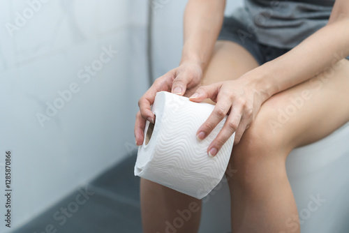 Fotografie Stomach ache, sick asian young woman holding stomach pain and holding tissue paper roll in bathroom sitting on toilet bowl, girl suffer from diarrhea her stomach painful