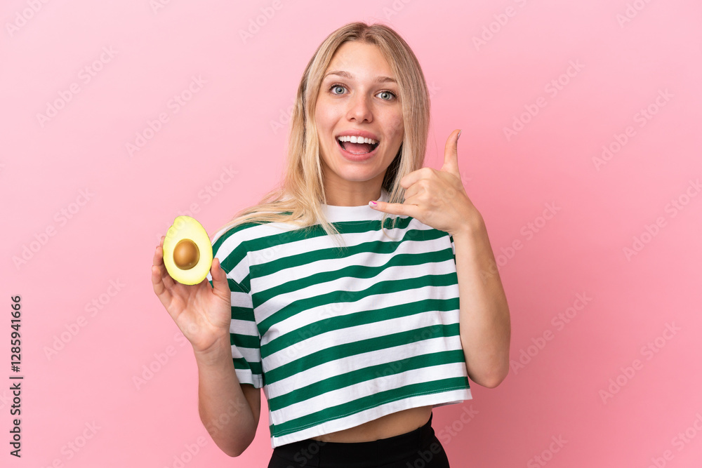 Young caucasian woman holding an avocado isolated on pink background making phone gesture. Call me back sign Young caucasian woman holding an avocado isolated on pink background making phone gesture. Call me back sign