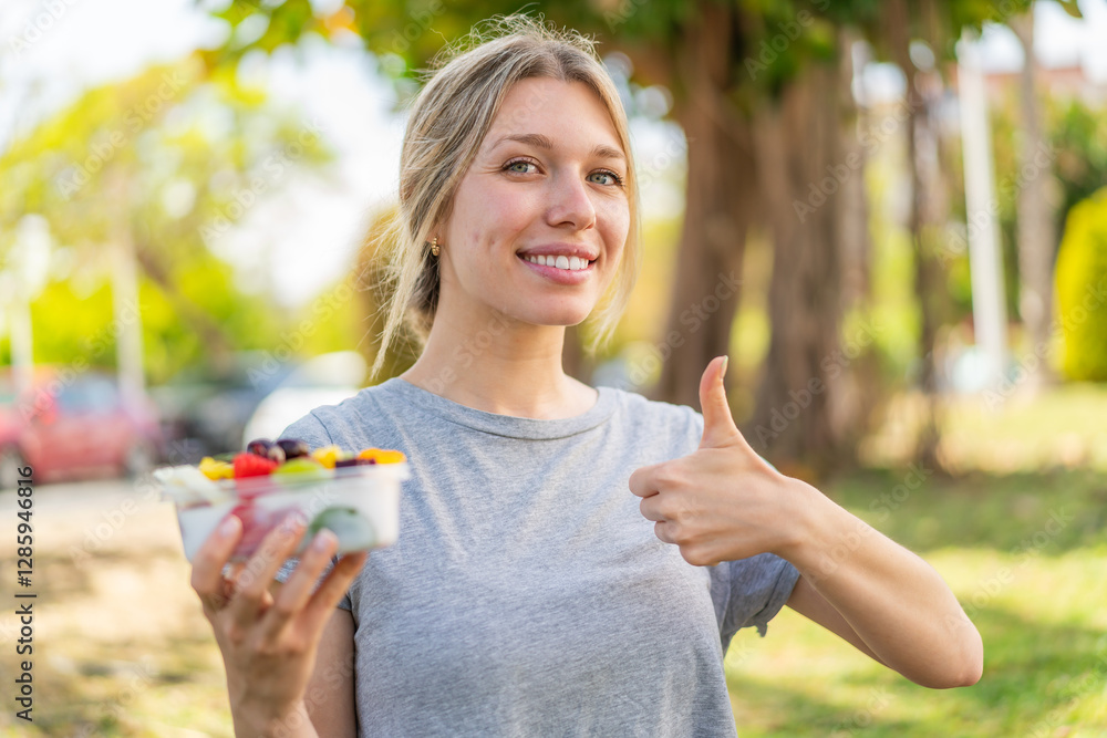 Young blonde woman holding a bowl of fruit at outdoors with thumbs up because something good has happened Young blonde woman holding a bowl of fruit at outdoors with thumbs up because something good has happened