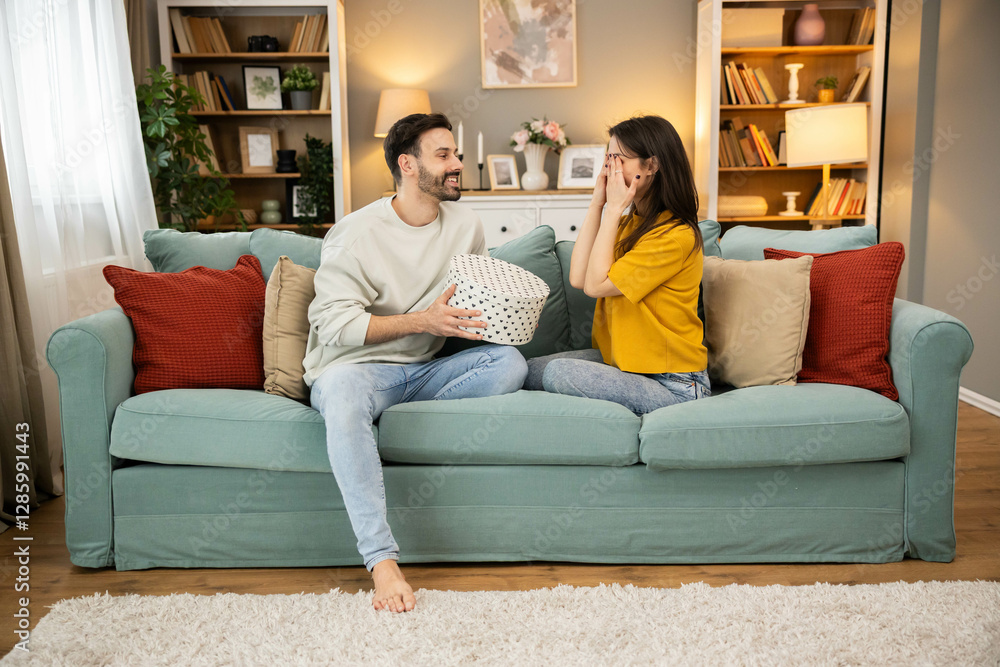Obraz premium A woman is playfully surprised at her birthday gift while her partner shares the joy on a comfy couch in a cozy living room.