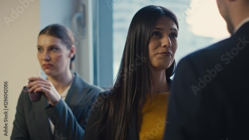 Professional woman in a suit holding a coffee cup observes a conversation between colleagues with a displeased expression. Concept of workplace dynamics, jealousy, and office rivalry
