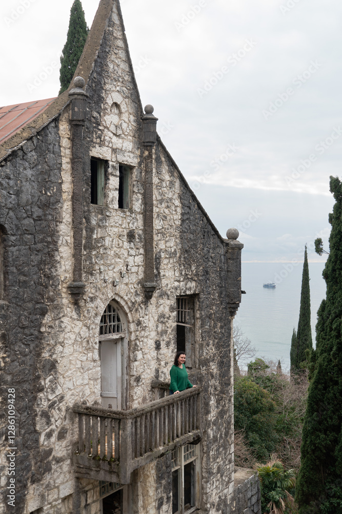 Fototapeta premium A woman in a green outfit stands on a stone balcony of an abandoned house in Gagra, Abkhazia