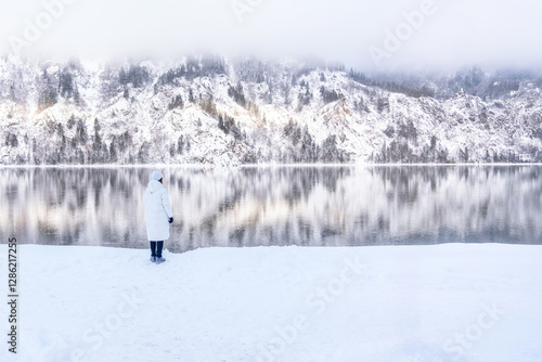 A small woman in a white jacket looks to the right at the majestic snow-capped mountains and their reflection in the Yenisei River near the city of Divnogorsk in winter in Russia photo