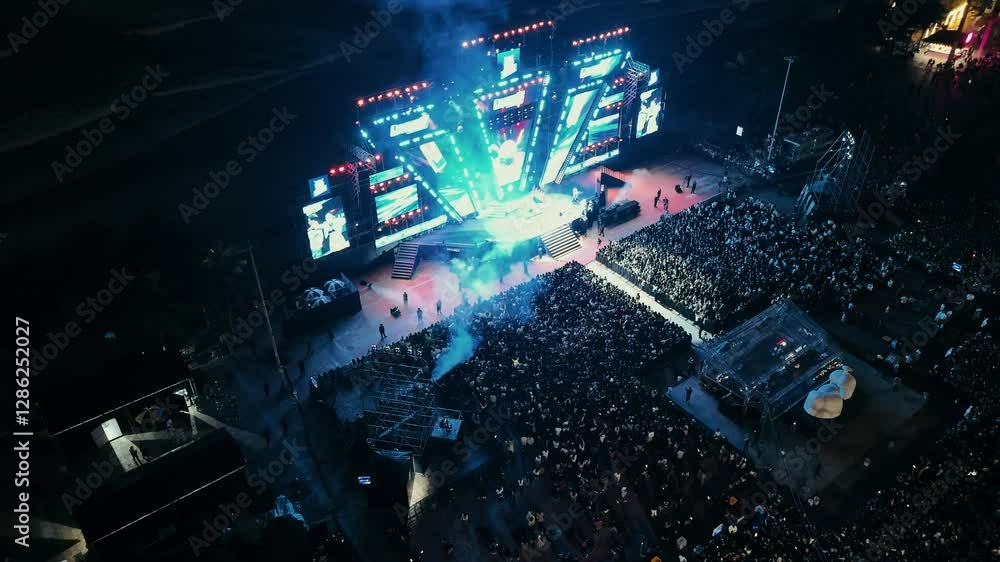 Open air music concert festival. Massive crowd gathering for night event near coastline. Colorful stage lighting illuminating energetic music performance with dynamic visual effects