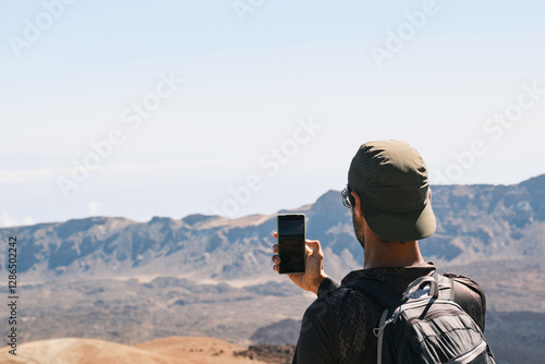 Man with a backpack takes a photo of a stunning mountain landscape with his smartphone.He captures the vastness of the desert scenery on a sunny day.Ideal for travel, adventure, and exploration themes