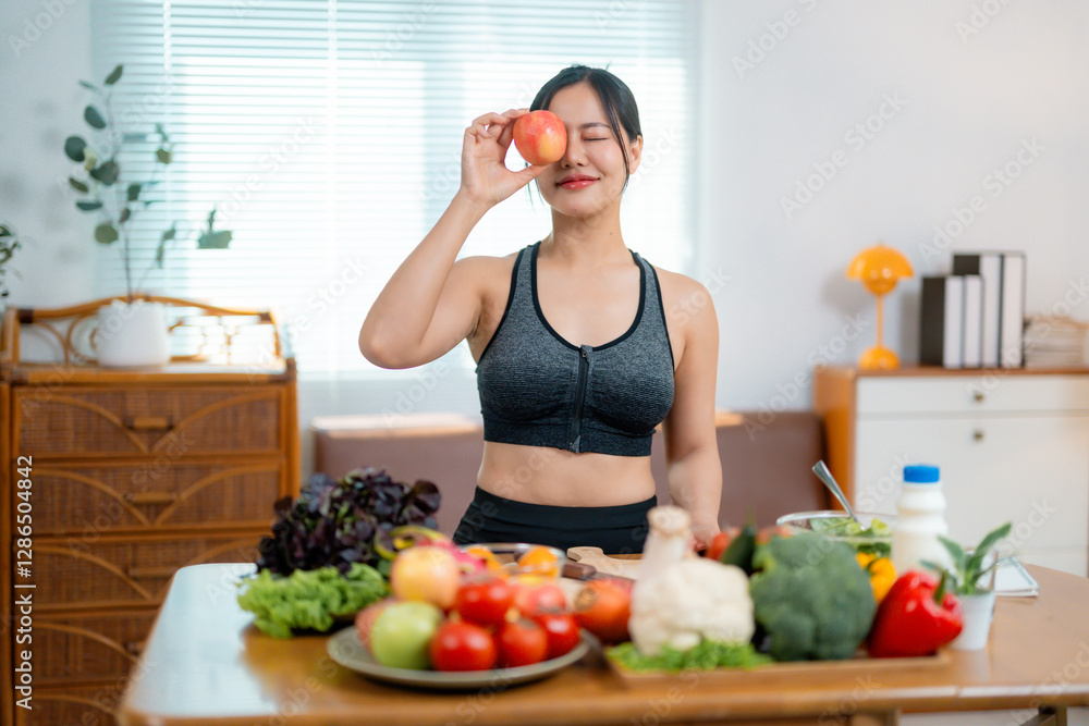 Sporty female blocking eye with red apple, posing near table displaying vibrant produce. Cheerful demeanor highlighting nutritious food selection and wellness approach Sporty female blocking eye with red apple, posing near table displaying vibrant produce. Cheerful demeanor highlighting nutritious food selection and wellness approach