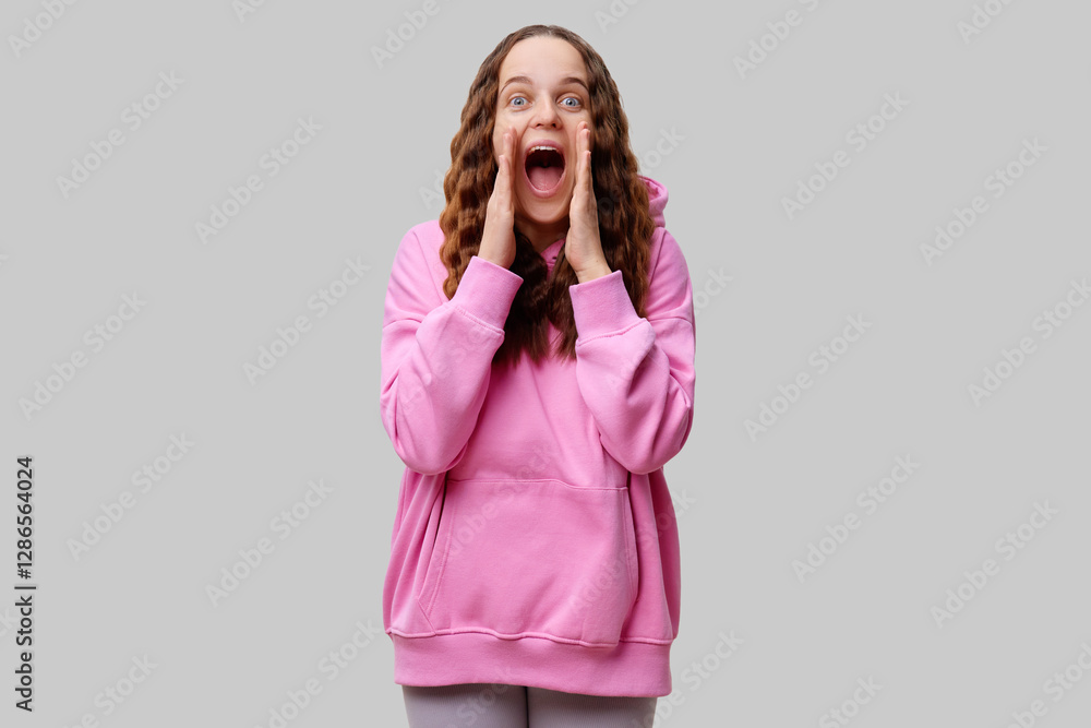 A young woman with long, wavy hair wears a pink hoodie and shows excitement by dramatically opening her mouth and raising her hands to her face. Her expression conveys pure joy A young woman with long, wavy hair wears a pink hoodie and shows excitement by dramatically opening her mouth and raising her hands to her face. Her expression conveys pure joy