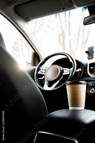 A recyclable coffee cup and lid are on the armrest next to the driver in the car interior, view from the back seat of the car. Coffee in the car. Sun rays in the car. Traveling in the car with coffee 