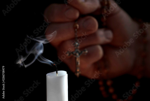 Blown out white candle with smoke in front of male hand with rosary on black background, the extinguished candle symbolizes the last prayer for a deceased person and belief in life after death