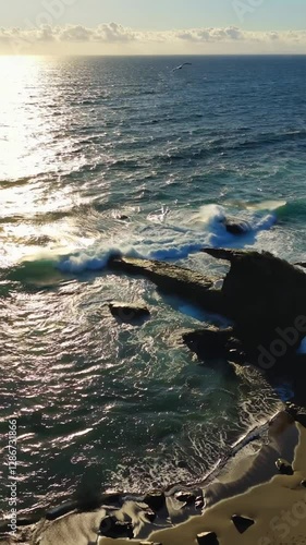 Aerial view of waves breaking over rocks in ocean. Ocean waves in beautiful aerial view of beach drone shot. Bird's eye view of ocean waves crashing against an empty rocky stone cliff from above