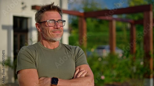 Portrait of confident successful man smiling with crossed arms, enjoying garden and morning sunlight at home. Mature, middle-aged happy male in glasses smiling outdoor. Fit and healthy senior.