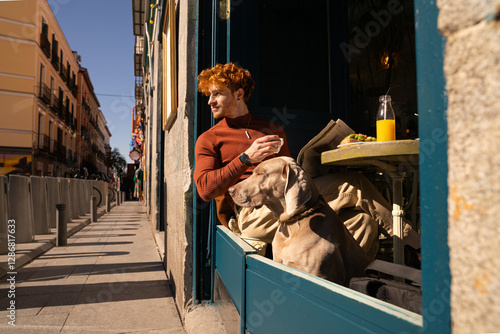 Sunny scene of Young ginger haired boy and his dog on the terrace of a cafe having a healthy breakfast