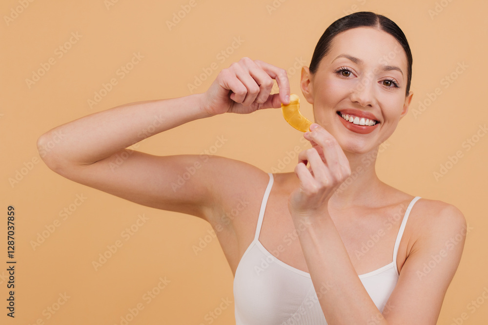 Smiling woman holding golden eye patches for skincare treatment on beige background Smiling woman holding golden eye patches for skincare treatment on beige background