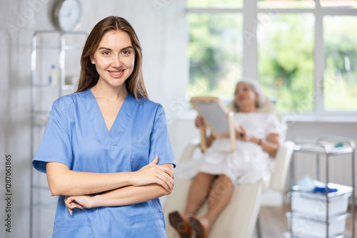 Smiling young lady wearing medical scrub standing at treatment room in front of female patient lying at examination couch. photo