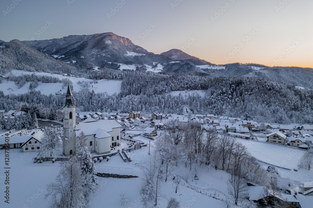 Beautiful church at Ljubno ob Savinji in cold winter morning. Visible haze from the river, sun rising up and smoke coming out of smokestacks.