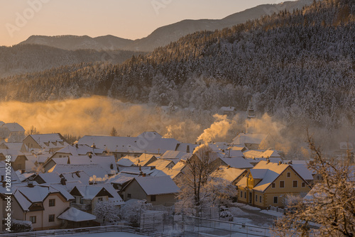 Obraz na płótnie Beautiful village of Ljubno ob Savinji in cold winter morning. Visible haze from the river, sun rising up and smoke coming out of smokestacks. w salonie